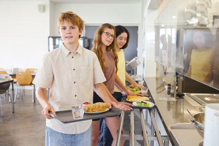 Students in a clean school cafeteria waiting in line to receive their meals, carrying trays with their selected food. The atmosphere is organized, bright, and emphasizes healthy eating and amicable interaction.の写真素材