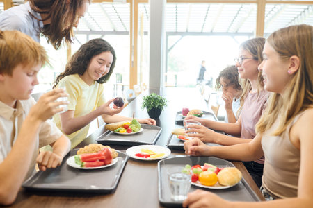 A group of schoolchildren having lunch in a clean cafeteria, enjoying nutritious meals together in a positive and friendly environment. The table is set with fresh fruits, colorful dishes, and beverages.の写真素材