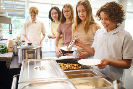 Diverse group of students queuing for lunch in a well-lit school cafeteria, enjoying freshly prepared food and friendly interactions, emphasizing a positive and healthy eating experience in a clean and inviting setting.の写真素材