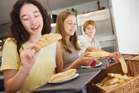 Three students pass through the school cafeteria, choosing fresh bread and other items. The scene portrays a cheerful environment, emphasizing teamwork, nutritious meals, and social interaction during lunchtime in a school setting.の写真素材