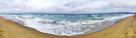 Panoramic view of a sandy beach with ocean waves meeting the shore and soft cloudy sky.の写真素材