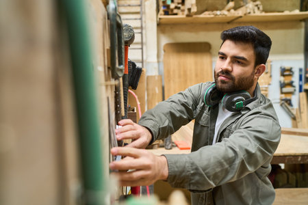 A young man wearing headphones operates machinery in a lumberyard. He is focused and wearing a work...