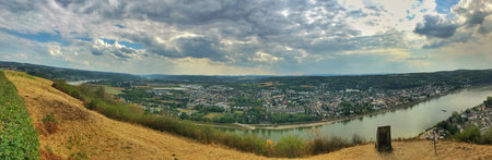 Wide panoramic view showcasing a scenic riverside town under a dramatic cloud-filled sky.
