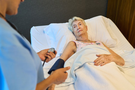 A healthcare professional checks the blood pressure of an elderly patient in a rehabilitation center. The patient lies in bed, receiving attentive care from an experienced and compassionate medical staff.の写真素材