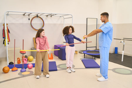 A friendly pediatric physiotherapy session where a therapist supervises two young girls as they practice balance and stick exercises. Fun, safe rehabilitation environment focused on motor skills and movement.の写真素材