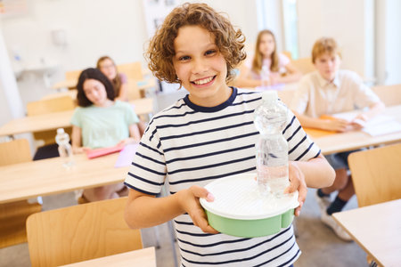 A cheerful child in casual attitude holding a meal tray and water bottle, surrounded by classmates in a bright, modern classroom promoting a supportive and engaging learning environment.の写真素材