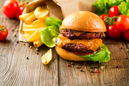 Hamburger Vintage style. Burger with two fried beef cutlets and fresh tomatoes with lettuce on wooden table. Cheeseburger served with French Fries in crumpled brown paper. Junk food conceptの写真素材
