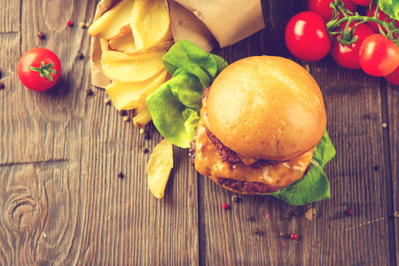 Tasty burger with fried juicy beef patties and fresh tomatoes with lettuce over wooden table. Hamburger with vegetables close-up. Cheeseburger with fried potatoes on rustic surface. Vintage styleの写真素材