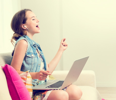 Beautiful teenage girl sitting on sofa at home, using laptop computer, laughing and having fun. Happy Teenage girl enjoying the music with headphones and dancing. Emotions. Happiness. Good moodの写真素材
