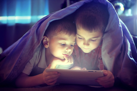 Two kids using tablet pc under blanket at night