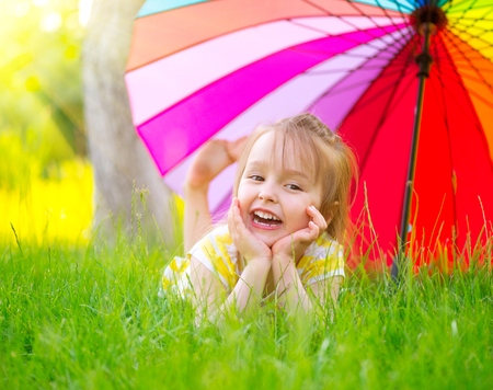 Portrait of a smiling little girl lying on green grass under the colorful umbrellaの写真素材