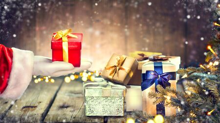 Santa Claus hand holding Christmas gift box over wooden background. Decorated Christmas tree with holiday giftsの写真素材