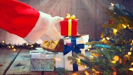 Santa Claus hand holding Christmas gift box over wooden background. Decorated Christmas tree with holiday giftsの写真素材