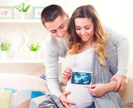 Happy young couple expecting baby. Beautiful pregnant woman and her husband together holding ultrasound picture of their babyの写真素材