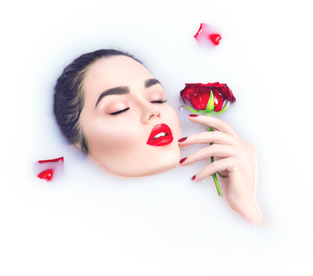 Beautiful model girl with bright makeup holding red rose flower in her hand and relaxing in milk bathの写真素材