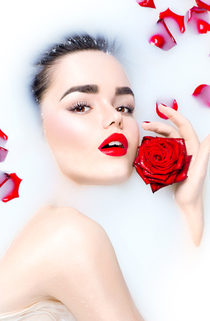 Beauty young model girl with bright makeup and red rose flower relaxing in milk bathの写真素材