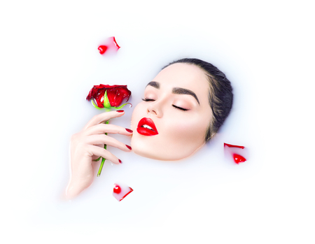 Beautiful model girl with bright makeup holding red rose flower in her hand and relaxing in milk bathの写真素材
