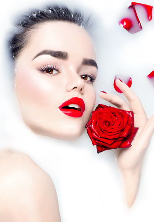 Beauty young model girl with bright makeup and red rose flower relaxing in milk bathの写真素材