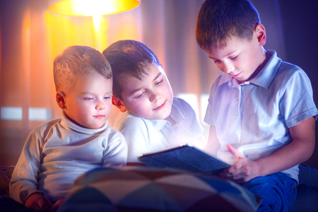 Three little boys with tablet computer in a dark room. Kids playing games on tablet pcの写真素材