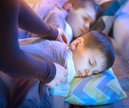 Children sleeping and dreaming in bed. Mother cover her little sons with a blanketの写真素材