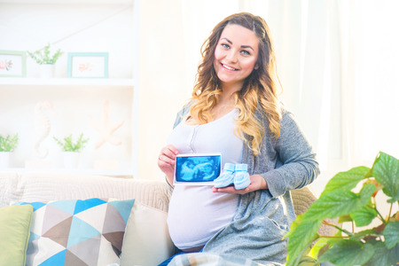 Pregnant happy woman holding blue baby shoes and ultrasound picture of her baby in her handsの写真素材