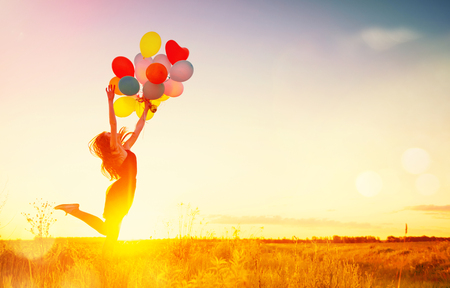 Beauty girl running and jumping on summer field with colorful air balloons over sunset skyの写真素材