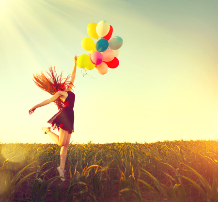 Beauty redhead girl running and jumping on summer field with colorful air balloons over clear blue skyの写真素材