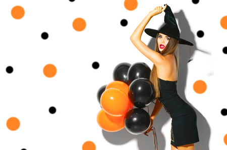 Halloween party girl. Sexy witch holding black and orange air balloons. Beautiful young woman in witches hat and costumeの写真素材