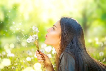 Beautiful young woman lying on green grass and blowing dandelions. Allergy free conceptの写真素材