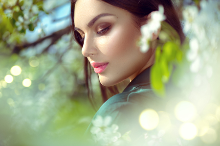 Beauty young woman enjoying nature in spring apple orchard, Happy Beautiful brunette girl in Garden with blooming apple trees. Smiling Person smelling blossom flowers outdoors. Fashion model portraitの写真素材