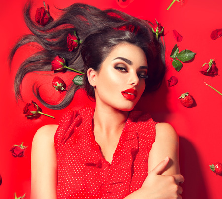 Beauty sexy model girl lying on red background with rose flowers and strawberries. Beautiful brunette young woman with long hair and perfect make-up, red seductive lips, smoky eyes. Trendy makeupの写真素材
