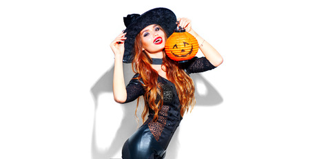 Halloween. Sexy witch with bright holiday makeup. Beautiful young woman posing in witches costume with pumpkin lantern over white backgroundの写真素材