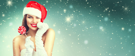 Christmas woman. Joyful model girl in Santa's hat with red lips and lollipop candy in her hand. Closeup portrait over winter snow backgroundの写真素材