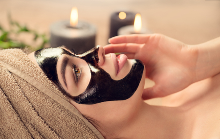Beautiful woman with black purifying black mask on her face. Beauty model girl with black facial peel-off mask lying in spa salon. Skin care, acne treatment, cleansing skin. Peel of charcoal maskの写真素材