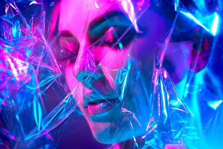 Fashion model woman in colorful bright neon lights posing in studio through transparent film. Portrait of beautiful sexy girl in UV. Art design colorful makeupの写真素材