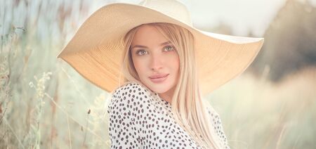 Beautiful model girl posing on a field, enjoying nature outdoors in wide brimmed straw hat. Beauty blonde young woman with long straight blond hair closeup portraitの写真素材