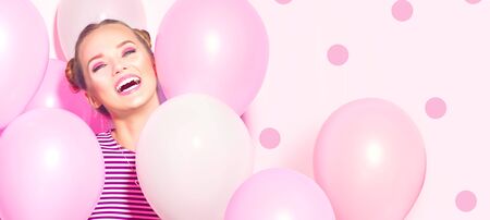 Beauty girl with colorful air balloons laughing over pink background. Beautiful Happy Young woman on birthday holiday party. Joyful model having fun, playing and celebrating with pastel color balloonの写真素材