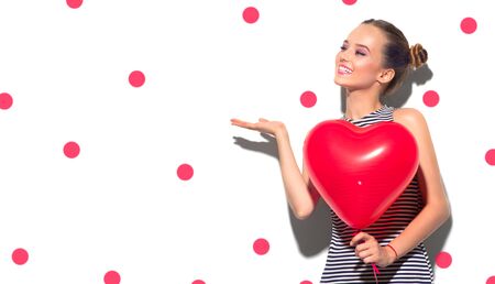 Valentine Beauty girl with red air balloon portrait pointing hand, isolated on white background. Beautiful Happy Young woman presenting products. Holiday party, birthday. Valentines's Day celebratingの写真素材