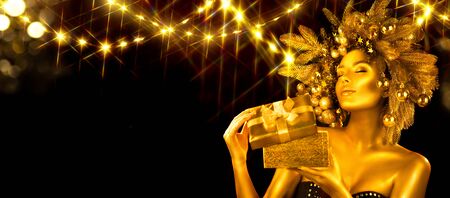 Christmas golden Woman. Winter girl receiving gift, opening magic box,  Beautiful New Year, Christmas Tree Holiday Hairstyle and gold skin Makeup. Girl in decorated Xmas wreath. Beauty surprised Modelの写真素材