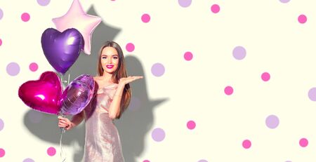 Valentine Beauty girl with pink heart shaped air balloons portrait pointing hand, isolated on polka dots background. Beautiful Happy Young woman presenting products. Holiday party, birthday.の写真素材