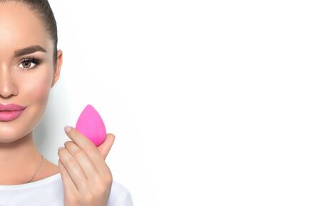 Beauty model girl, makeup artist holding pink make up blender sponge and smiling. Beautiful brunette young woman with nude make-up. Perfect skin closeup. Face contouring makeup. On whiteの写真素材