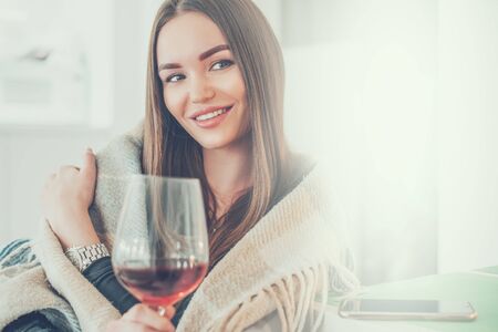 Beautiful model fashion girl drinking red wine at home and smiling. Beauty woman covered with plaid, holding wine glass in a cozy room. Relaxing femaleの写真素材