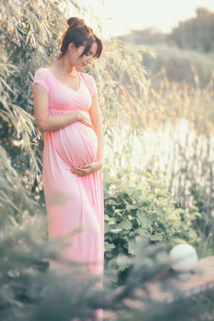 Pregnant happy Woman touching her belly. Full length Pregnant beauty young mother outdoor portrait, caressing her belly and smiling. Healthy Pregnancy concept, brunette expectant female in summer parkの写真素材