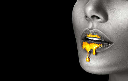 Lipstick dripping. Paint drips, lipgloss dripping from sexy lips, liquid Gold metallic paint drops on beautiful model girl's mouth, creative make-up. Desaturated Beauty woman face makeup close up. Artの写真素材