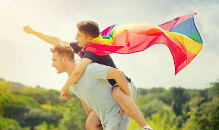 Happy young couple, gay family with flying LGBT rainbow flag having fun outdoors. Joyful gay men together in a park. laughing, hugging, enjoying nature outside, over sunny sky. Super hero. Piggyback.の写真素材