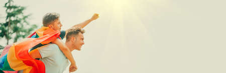 Happy young couple, gay family with flying LGBT rainbow flag having fun outdoors. Joyful gay men together in a park. laughing, hugging, enjoying nature outside, over sunny sky. Super hero. Piggybackの写真素材