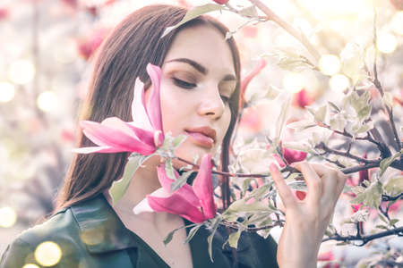 Beauty young woman enjoying nature in spring spring magnolia flowers. Beautiful brunette girl in Garden with blooming magnolia trees. Smiling girl with blossom flowers outdoors. Fashion model portraitの写真素材