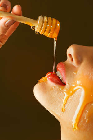 Honey dripping from honey dipper on sexy girl lips. Thick honey dipping from the wooden honey spoon. Beauty model woman open mouth, model eating nectar. Healthy food concept, diet,の写真素材