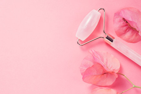 Face Jade roller over pink background with flowers. Facial lifting massage tool, anti aging treatment, gua sha pink quartz rolling device for skin care, self massaging accessory. Bの写真素材