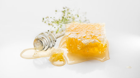 Fresh Honey in honeycombs dripping, pouring from a glass honey dipper spoon, on grey background. Honeycomb close-up. Healthy organic liquid honey spill, with flowersの写真素材
